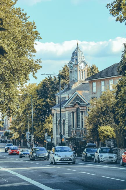 The image shows a busy street scene with several cars lined up in traffic, set in front of a historic church building with a prominent clock tower topped by a spire. The church features classic archit