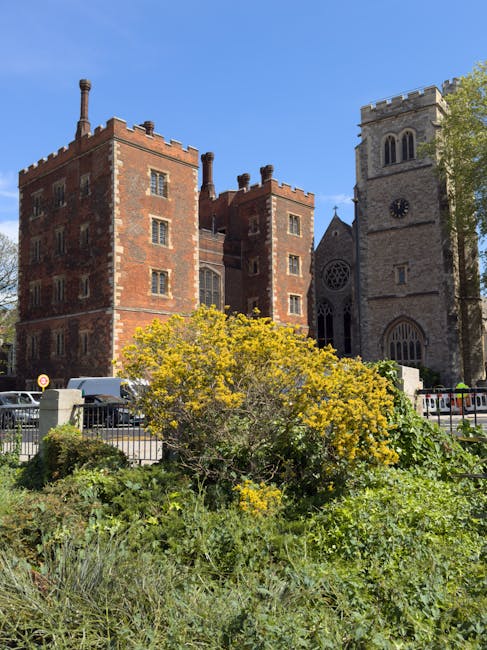 A vibrant bouquet arrangement of fresh flowers in shades of yellow, white, and green, featuring daisy-like blooms, roses, and filler foliage, held by a person in front of a historic brick building wit