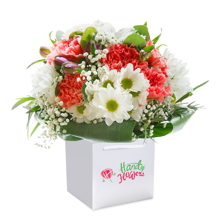 A meticulously arranged bouquet radiates from the centre of a crisp, square white gift bag, its striking presence perfectly suited to the refined tastes of Shooter's Hill. Coral and peach carnations, lavish with ruffled petals that seem almost to flutter in the light, draw the eye as the bouquet's heart. Clustered between them are luminous white daisies, their golden-green eyes exuding a playful brightness reminiscent of the nearby Oxleas Meadow in full bloom. Wisps of delicate white gypsophila are lightly scattered throughout, enhancing the arrangement's airy, almost weightless feel. Unfurling buds tinged with a hint of purple add subtle intrigue and depth to the left edge, satisfying the eye's search for gentle surprises. Encircling these lively blooms, an abundance of rich, deep green foliage and broad, lush leaves build a sturdy, natural base, evoking the canopy of mature local trees. The flowers are artfully shaped into a full, rounded form, a testament to the care and precision found in the best florists of Shooter's Hill, and the overall effect is one of generosity and freshness. The polished white gift bag, fitted with strong ribbon handles, hints at easy transport for any sender in SE18. This image, imbued with both elegance and the cheerful optimism of spring, serves as a beautiful emblem for the best in local floral delivery-perfect for birthdays, anniversaries, or simply sharing a heartfelt message with a neighbour.