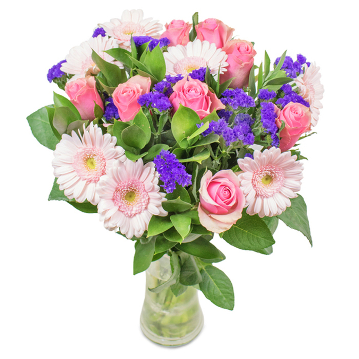 This radiant Shooter's Hill bouquet exudes refined cheer-a masterful composition arranged in a clear glass vase and set against a pristine backdrop, its colours and textures reflecting the nuanced artistry of the local florist. At its centre, approximately seven to nine pink roses display a variety of blush and medium pink hues; their gently furled or freshly opened petals form classic spirals, creating a focal point of traditional romance. These are gracefully surrounded by five to seven pale pink Gerbera daisies, easily identified by their generous round blooms and delicately feathered petals surrounding subtle creamy centres. Sprinkled throughout are vivid clusters of purple statice, their crisp, textural appeal providing lively contrast that draws the eye and adds visual rhythm-much like the patchwork of bluebells and wildflowers dotting nearby woodland trails. The bouquet is anchored with lush, deep green foliage, whose abundant presence evokes the freshness of nearby commons on a spring day. The slender, transparent vase beautifully displays the intertwining stems and clear water, suggesting freshness and careful handling typical of a bespoke gift from a local Shooter's Hill florist. Suitable for a variety of occasions-from heartfelt congratulations on a new venture to birthday celebrations or a simple gesture of affection-this arrangement radiates joy, gentle sophistication, and a sense of homegrown abundance that is always present in this vibrant London community.